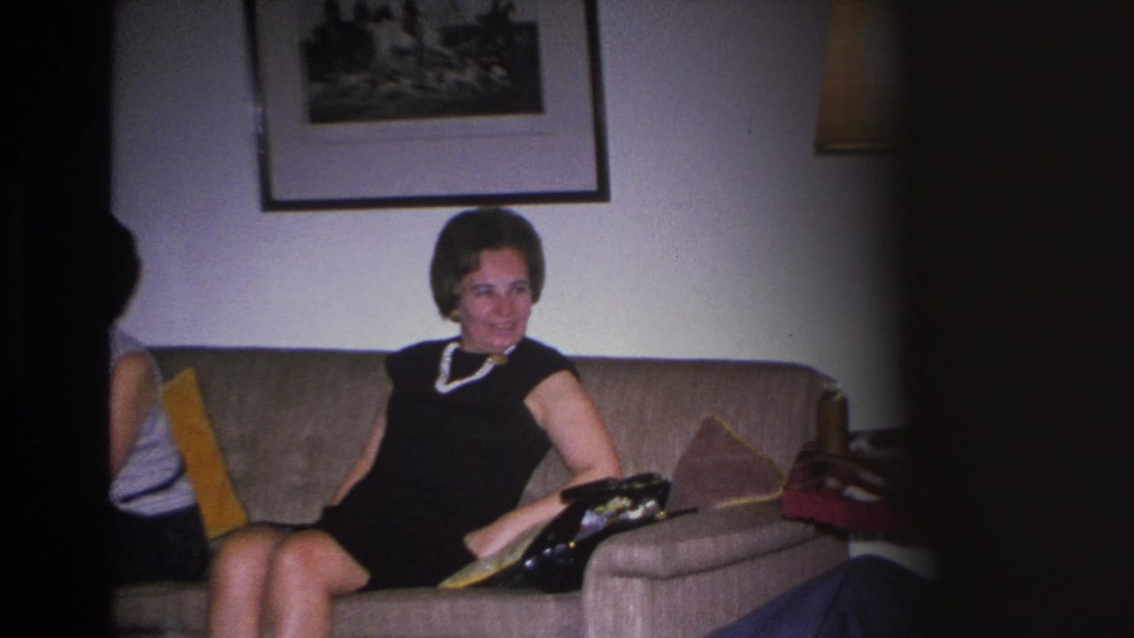 Woman in black dress and pearl necklace seated on a beige sofa in a 1960s American living room, with framed wall prints and a second person partially visible at the frame edge.
