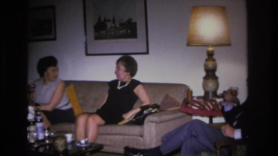 Wide shot of a 1960s American living room showing two women on a sofa, a lit drum-shade table lamp, and framed prints; a man's legs are partially visible at lower right.