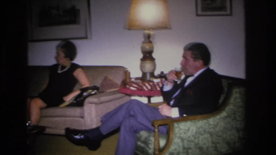 A woman in a black dress and pearl necklace on a sofa and a man in a dark suit holding a drink in an armchair in a mid-century American living room, circa 1966.