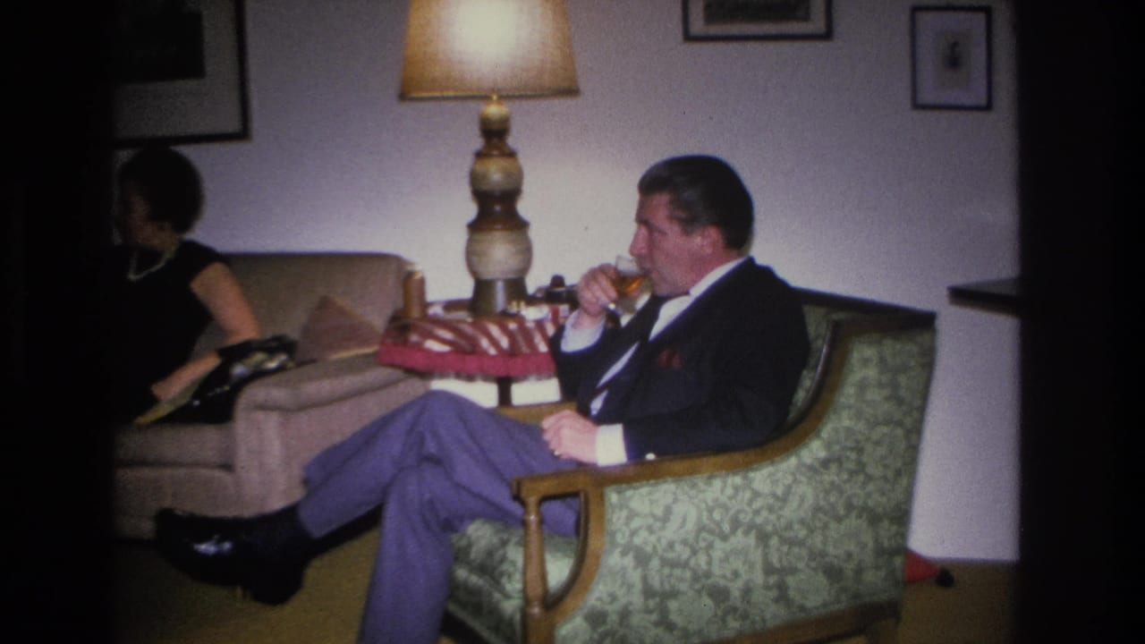 Man in 1960s dress seated in a green floral armchair drinking from a glass in a mid-century living room; woman in dark clothing partially visible on sofa behind him, Hoboken, New Jersey, circa 1966.