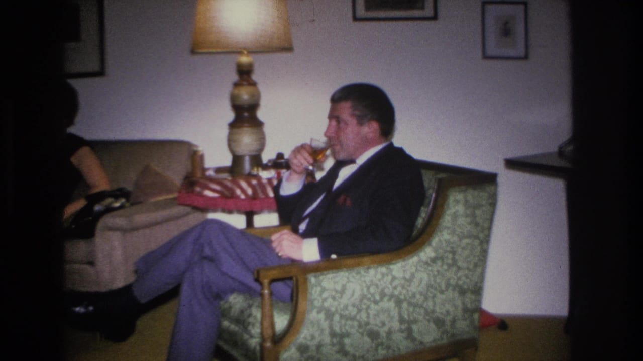 Man in mid-1960s dress seated in a green floral armchair raising a glass in a mid-century living room; woman in dark clothing partially visible at left, Hoboken, New Jersey, circa 1966.