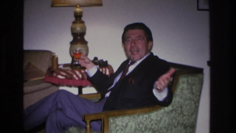 Close view of a man in a dark suit gesturing animatedly while holding a glass and appearing to speak or laugh in a mid-century living room interior, Hoboken, New Jersey, circa 1966.