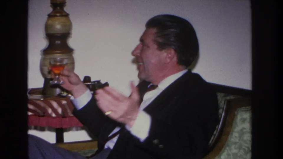 Man in a dark suit smiling broadly and gesturing with open hands while holding a glass in a mid-century living room interior, Hoboken, New Jersey, circa 1966.