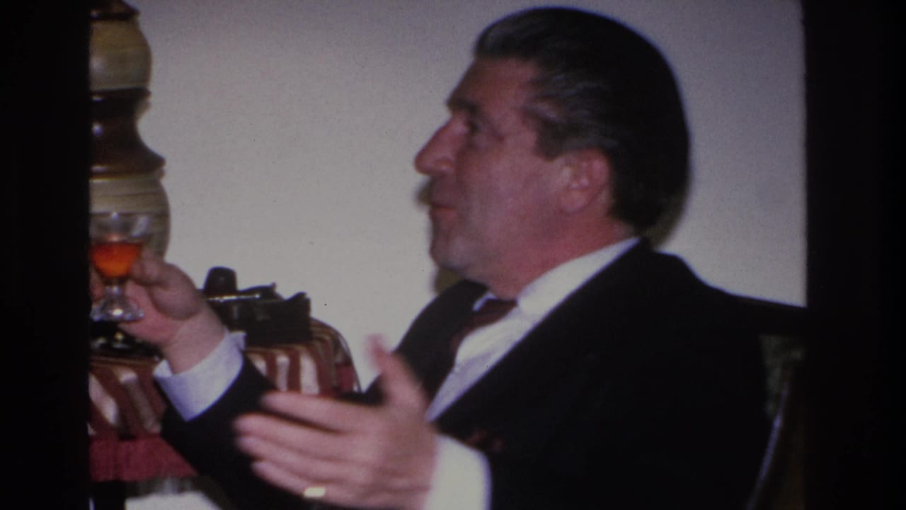 Middle-aged man in dark suit and tie holds a stemmed cocktail glass while gesturing mid-conversation, captured on 8mm at an indoor gathering, likely Hoboken, New Jersey, circa 1966.