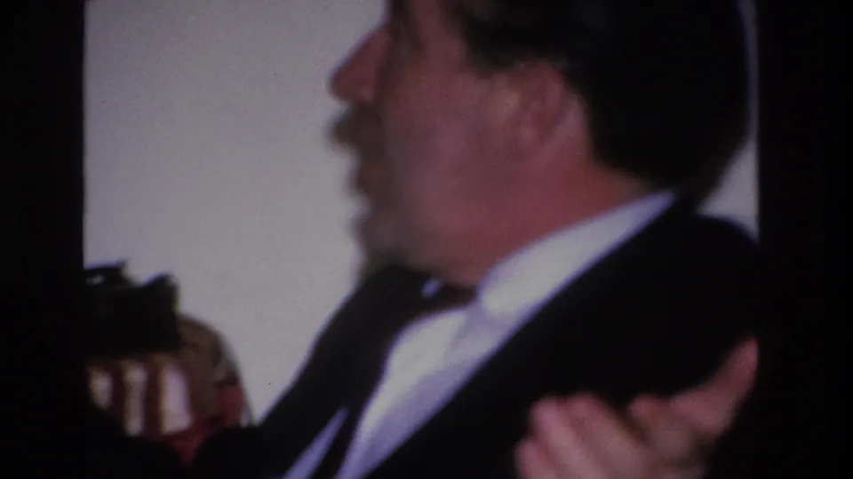 Same suited man gesturing mid-conversation at an indoor party; framed wall art visible in background, footage slightly out of focus, 8mm home movie circa 1966.
