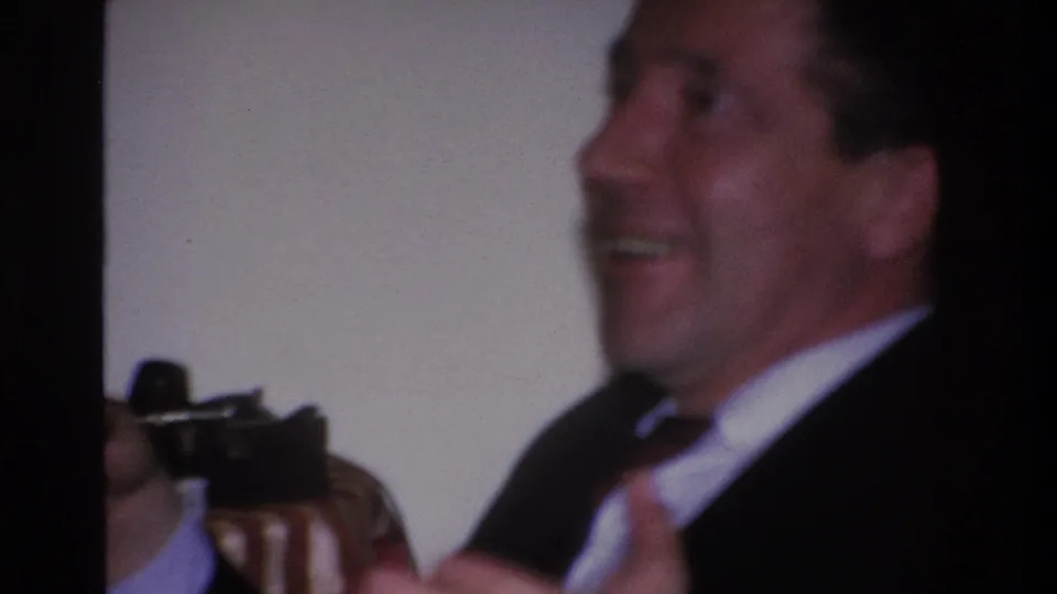 Close-up of man in dark suit and tie laughing mid-conversation at an indoor party circa 1966; a dark object in the lower left may be a second camera or party item.