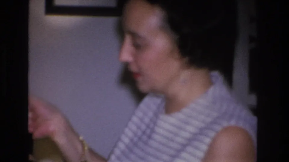 Woman in horizontal-striped sleeveless top gesturing mid-conversation at an indoor party; man in dark suit partially visible behind her, framed artwork on wall, 8mm home movie circa 1966.