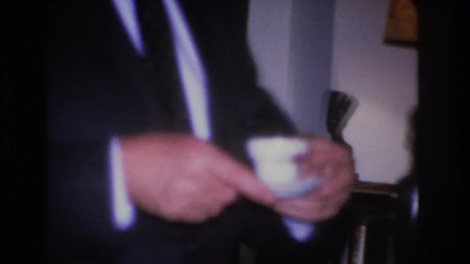 Man in a dark 1960s suit with white shirt and tie holds a small white teacup in what appears to be a mid-century domestic living room.