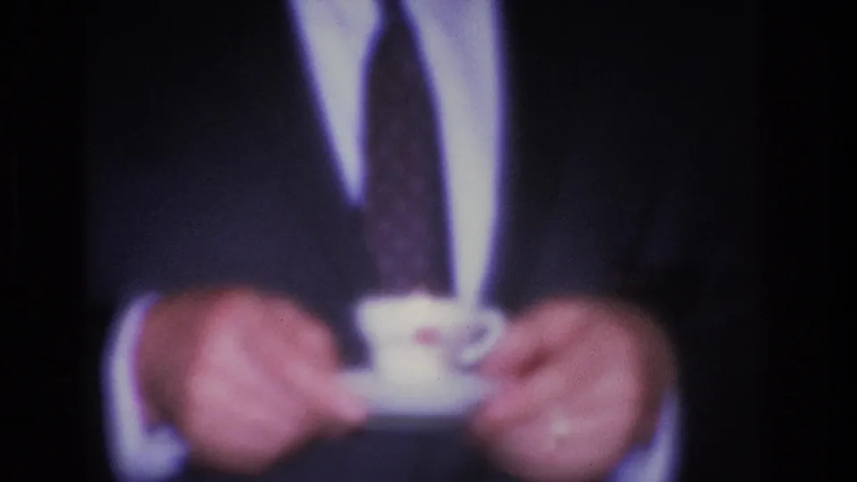Tight close-up of suited man's hands holding a white teacup and saucer; 8mm soft focus and grain visible in this domestic interior shot.
