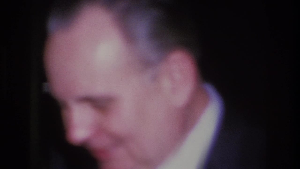 Three-quarter profile of a grey-haired man in a dark suit and tie, candid 8mm shot at a Hoboken, New Jersey social event, 1966.