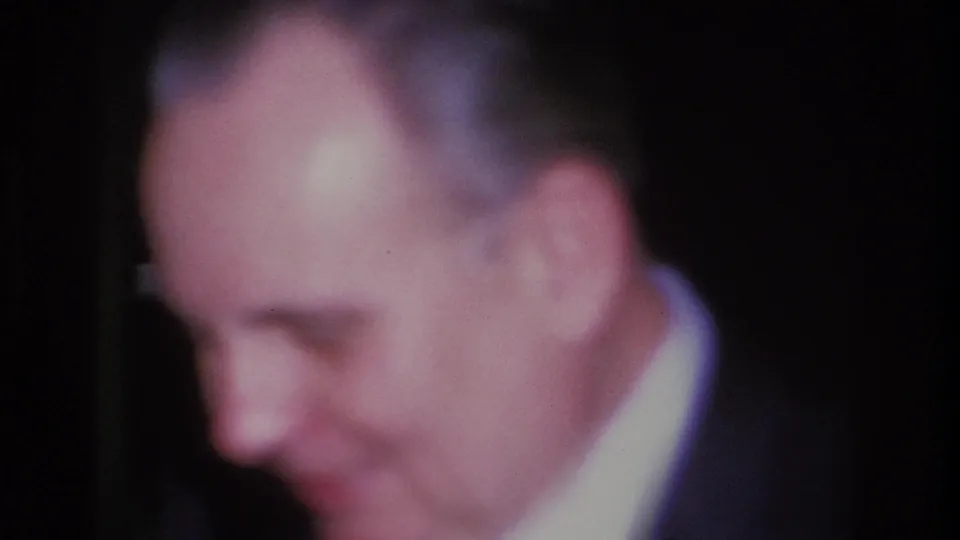 Three-quarter profile of a grey-haired man in a dark suit and tie, candid 8mm shot at a Hoboken, New Jersey social event, 1966.