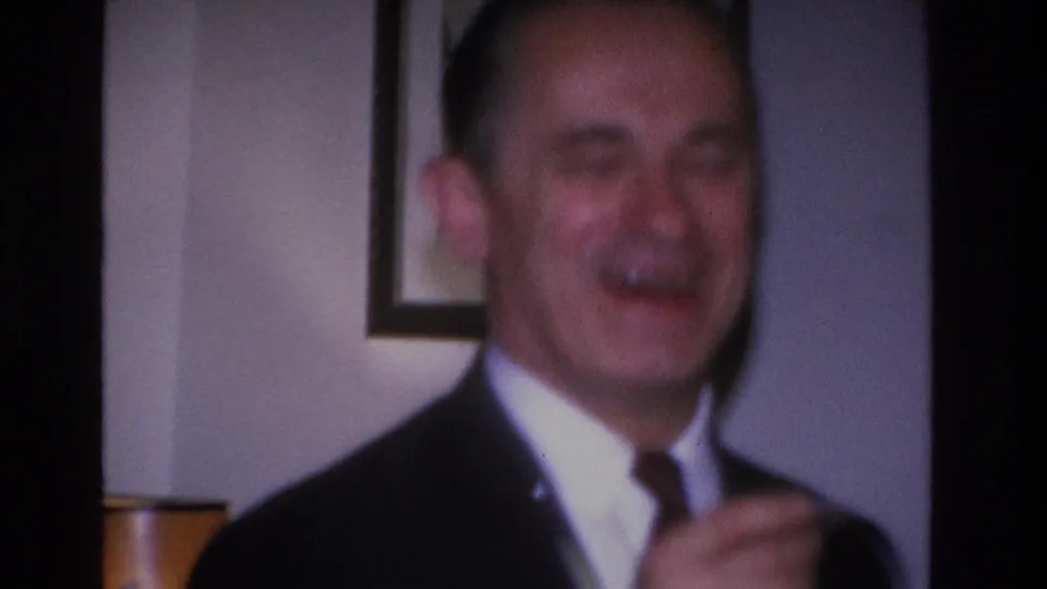Smiling man in a dark suit and tie at an interior social event, framed artwork on wall behind him, Hoboken, New Jersey, 1966.