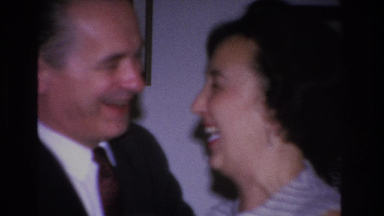 Man and woman in profile facing each other, both smiling, plain gray indoor background.