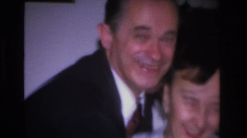 Man smiles directly toward camera in what appears to be a 1966 8mm home movie; woman in striped top visible at his side.