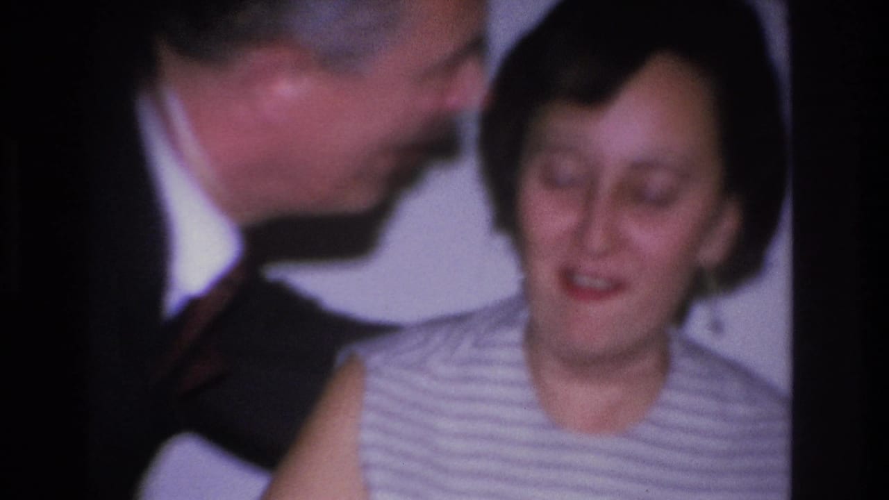 Close-up 8mm home movie frame from 1966 showing a man in a dark suit kissing a woman's cheek at a domestic gathering in Hoboken, New Jersey.