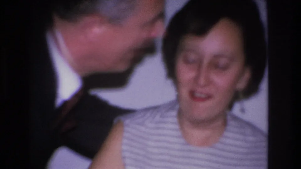 Close-up 8mm home movie frame from 1966 showing a man in a dark suit kissing a woman's cheek at a domestic gathering in Hoboken, New Jersey.