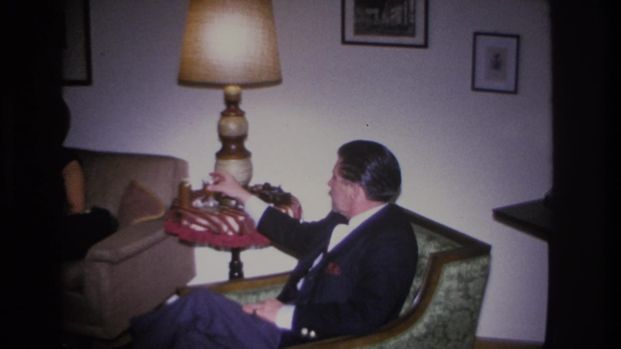 Wide living room shot from a 1966 home movie showing a man in a dark suit in a green armchair holding a red object, with a turned-wood floor lamp and framed prints on white walls.
