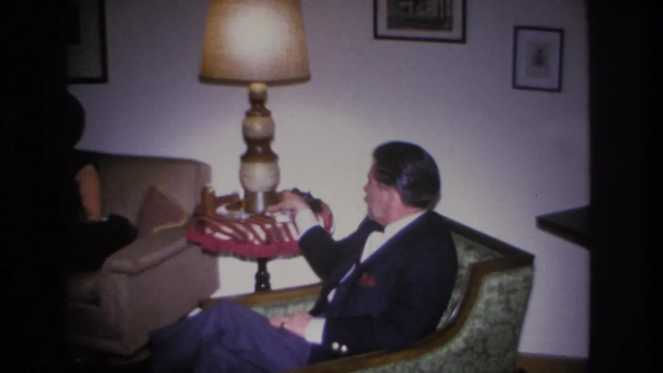 8mm living room frame from a 1966 home movie showing a man seated in a green armchair holding a red item, with a period floor lamp and framed wall art visible.