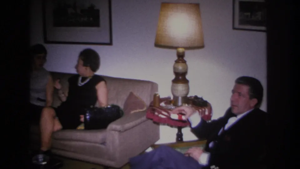 Wide 1966 home movie shot of a domestic gathering in Hoboken, New Jersey, showing two women on a sofa and a man in an armchair with period lamp and wall decor.