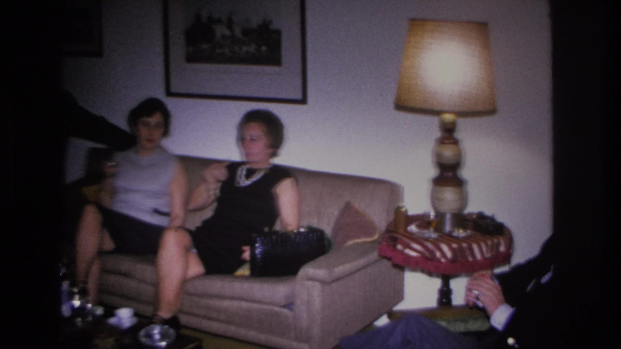Two women in 1960s attire, one in a black dress with pearls and one in a sleeveless gray top, seated on a sofa at a home party, Hoboken, New Jersey, circa 1966.