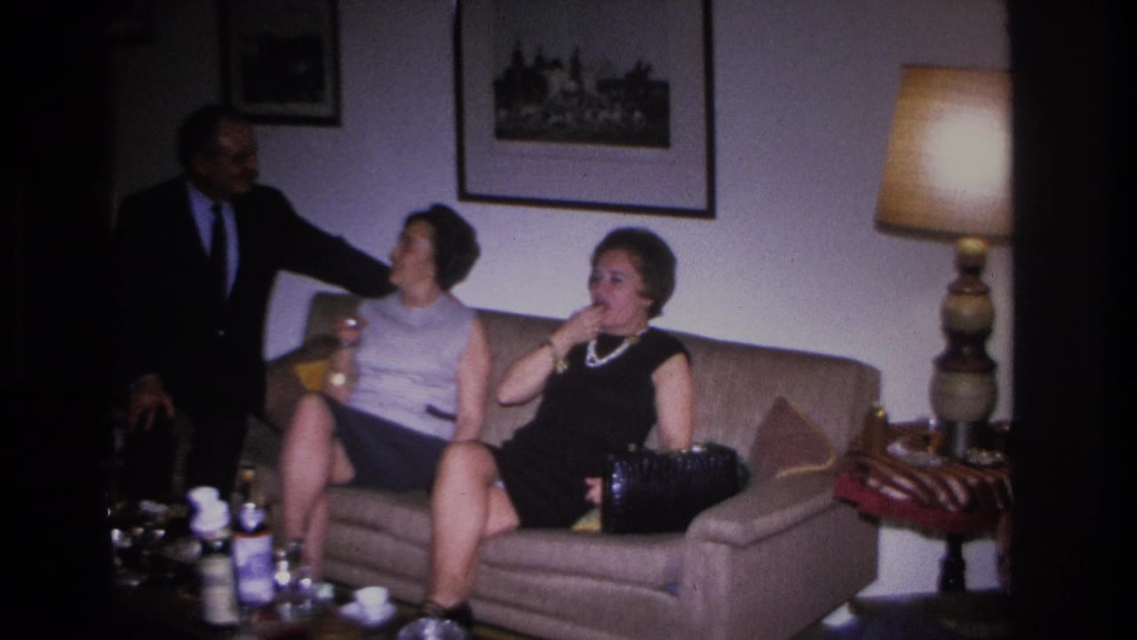 Man in dark suit stands beside two women on a sofa; living room lamp and framed art visible, bottles on the table.