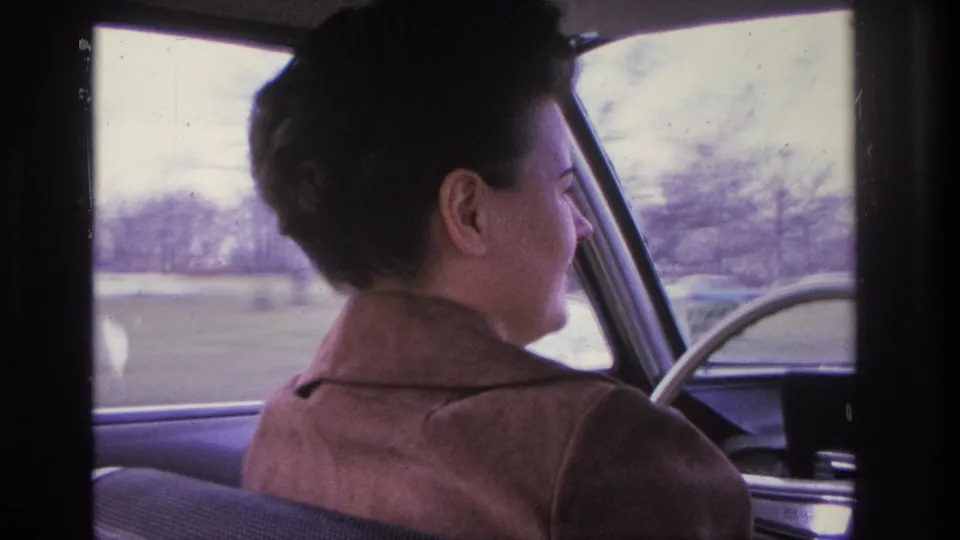 A woman in a brown jacket drives a vintage American car through a leafless suburban landscape, viewed from the rear seat, circa 1966.