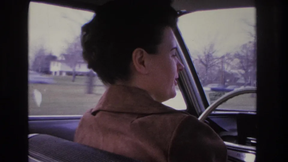 Close rear-seat view of a woman driving a vintage car through a bare-tree suburban street, a house visible in the background, circa 1966.