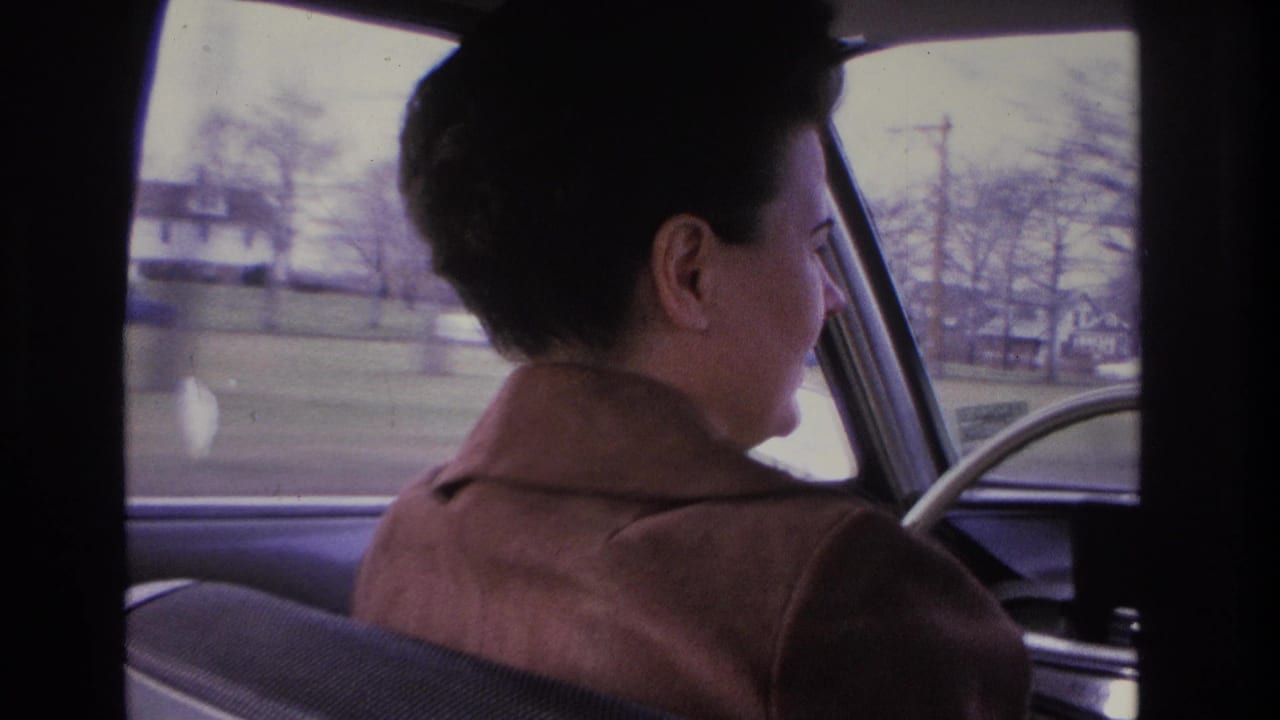 Back-seat view of a woman in a brown tailored coat driving a mid-1960s American car through a suburban residential neighborhood, possibly Hoboken, New Jersey, circa 1966.