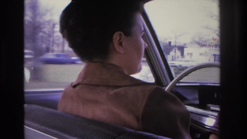 Back-seat view of a woman driving through a flat suburban neighborhood; a period blue automobile and low residential buildings visible through the window, circa 1966.