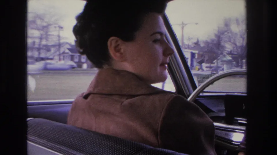 Woman turning toward the camera while driving; residential homes, utility poles, and a passing period automobile visible through the rear window, circa 1966.