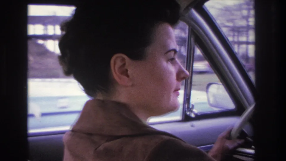 Partial profile of a woman turning at the steering wheel, identified as Hoboken, New Jersey, circa 1966; passing suburban buildings and bare trees visible through the car window.