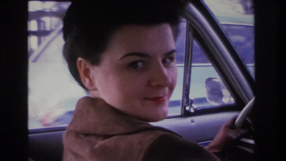 Woman smiling directly toward the camera over her shoulder; identified as a 1966 home movie shot in Hoboken, New Jersey; residential scenery visible through the rear window.