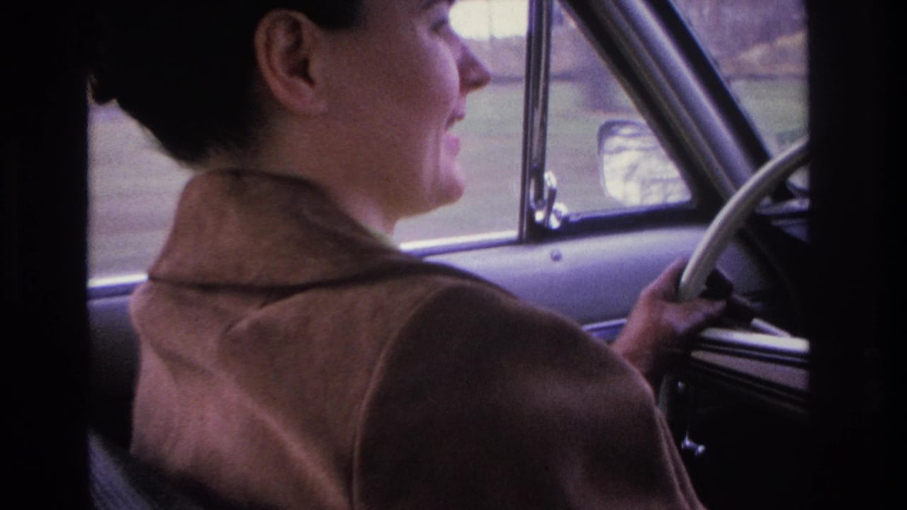 Woman from behind at the steering wheel of a classic car, right hand visible on the wheel rim.
