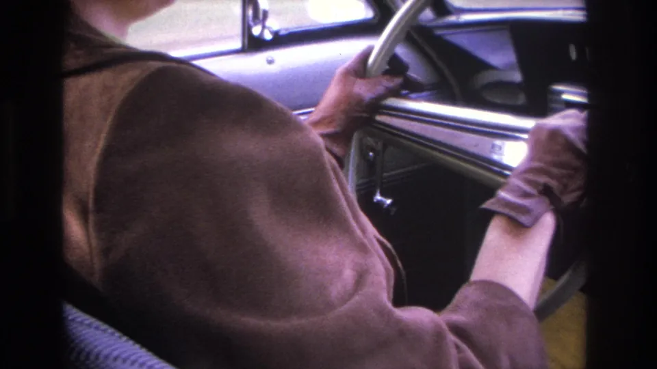 Close framing of a woman in a brown jacket at the steering wheel of a 1960s American car, showing chrome dashboard detail and yellow-toned fabric below.