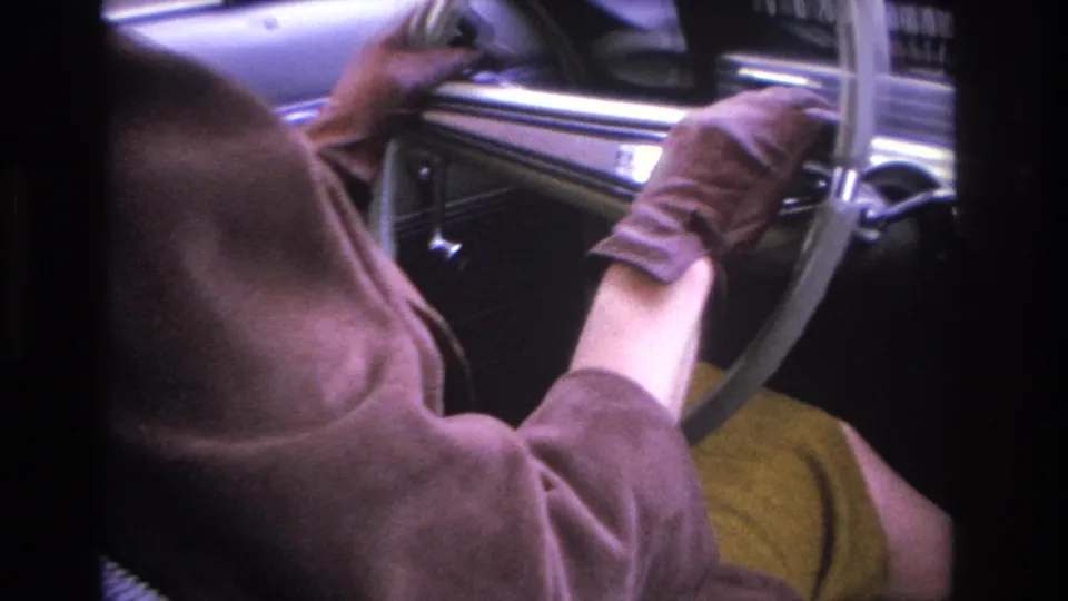 Close shot of hands gripping a chrome-rimmed steering wheel inside a 1960s American car, brown jacket visible, consistent with mid-1960s 8mm home-movie footage.