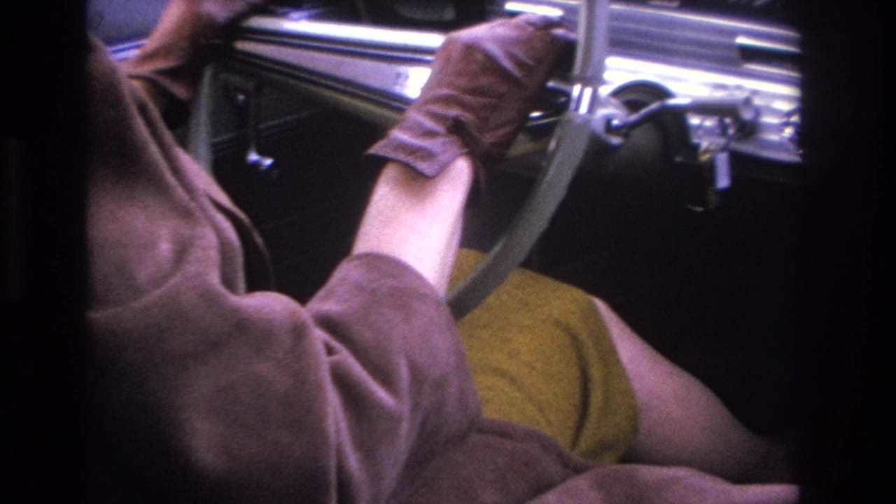 Close-up of a woman's torso in a brown coat and mustard skirt inside what appears to be a mid-1960s American car, gloved hands near the chrome instrument panel.