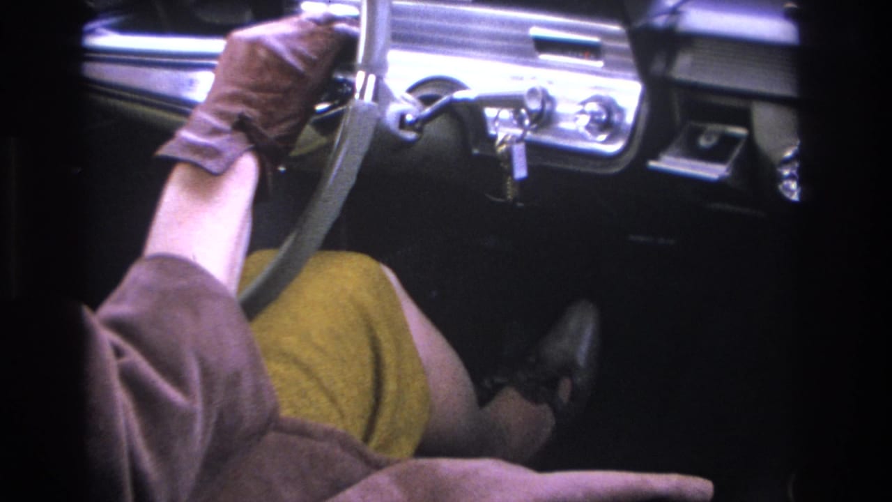 Woman's brown-gloved hands on a chrome-rimmed steering wheel inside a mid-1960s American car, mustard dress visible, circa 1966.