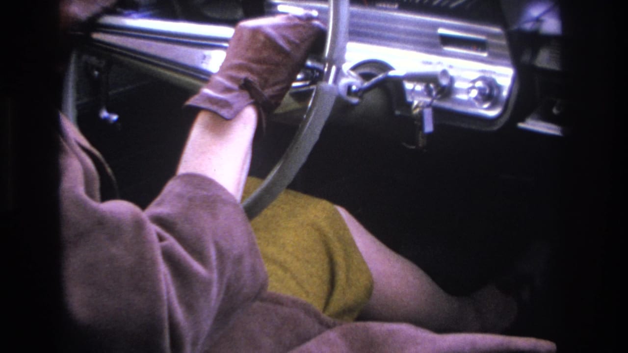 Woman's gloved hands on a chrome steering wheel; mustard dress and chrome dashboard details visible.