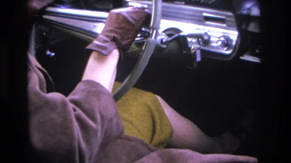 Brown-gloved hands gripping the steering wheel of a mid-1960s American automobile, mustard dress and chrome dash in view, circa 1966.