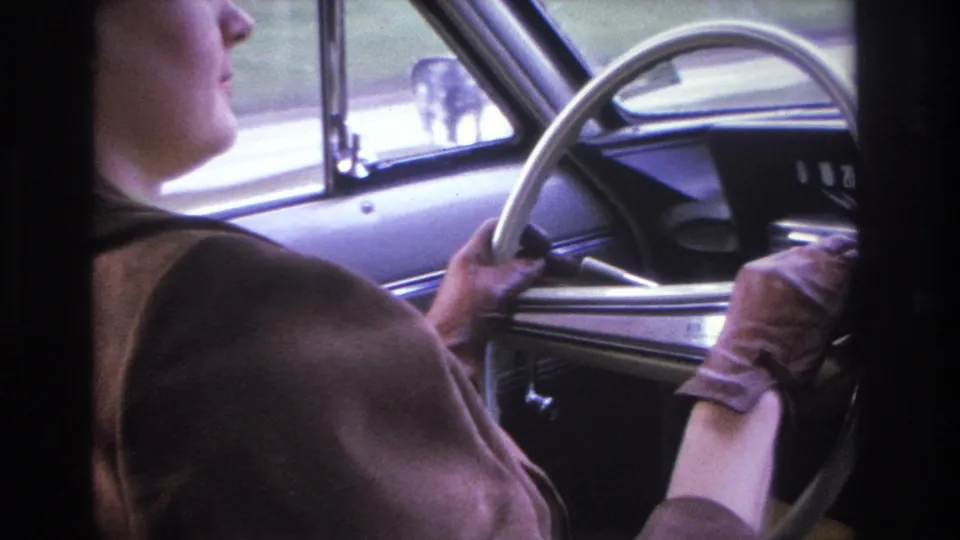 Partial profile of a woman in a brown coat at the wheel of a mid-1960s car; speedometer cluster and blurred road scene through the window, circa 1966.