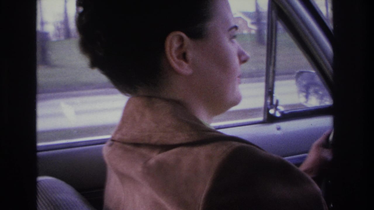 Woman driver with 1960s upswept hair and brown open-collar jacket at the wheel of a period American car, motion-blurred street visible through side window, Hoboken New Jersey area, circa 1966, 8mm film.