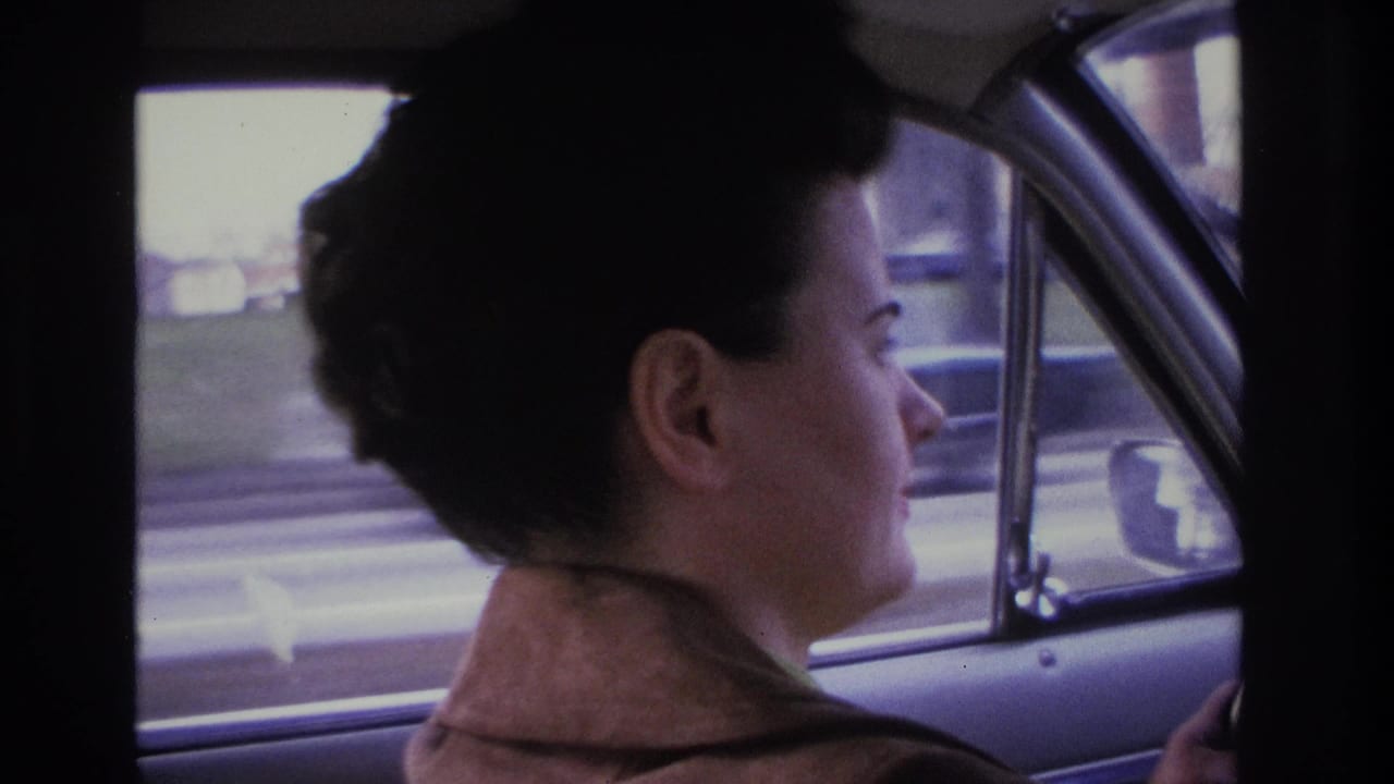 Woman driver with 1960s dark upswept hair and brown jacket, car window frame and chrome trim visible, blurred street scene with low commercial structures outside, circa 1966 8mm home movie.
