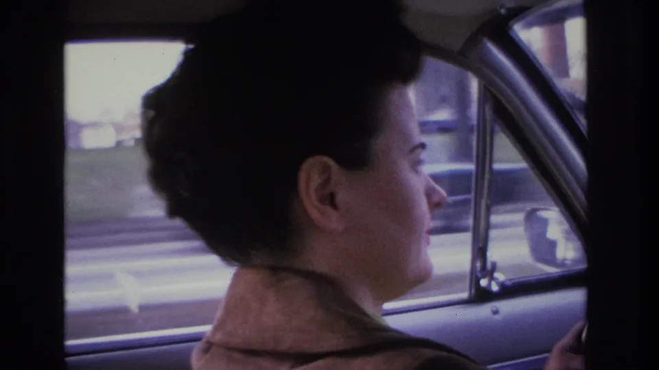 Woman driver with 1960s dark upswept hair and brown jacket, car window frame and chrome trim visible, blurred street scene with low commercial structures outside, circa 1966 8mm home movie.