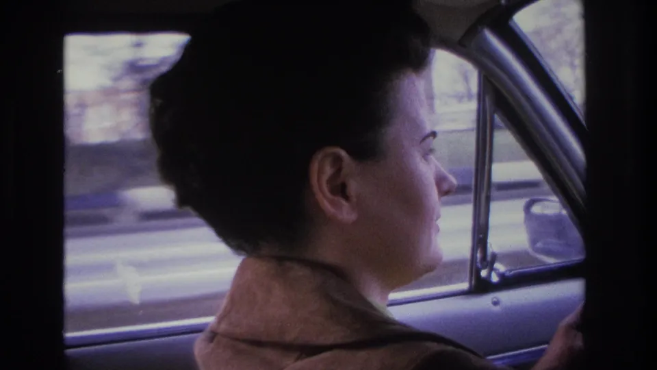 More frontal view of woman driving, dark 1960s upswept hair and brown jacket, car door interior panel visible at right edge, blurred street scenery outside window, 8mm home movie circa 1966.