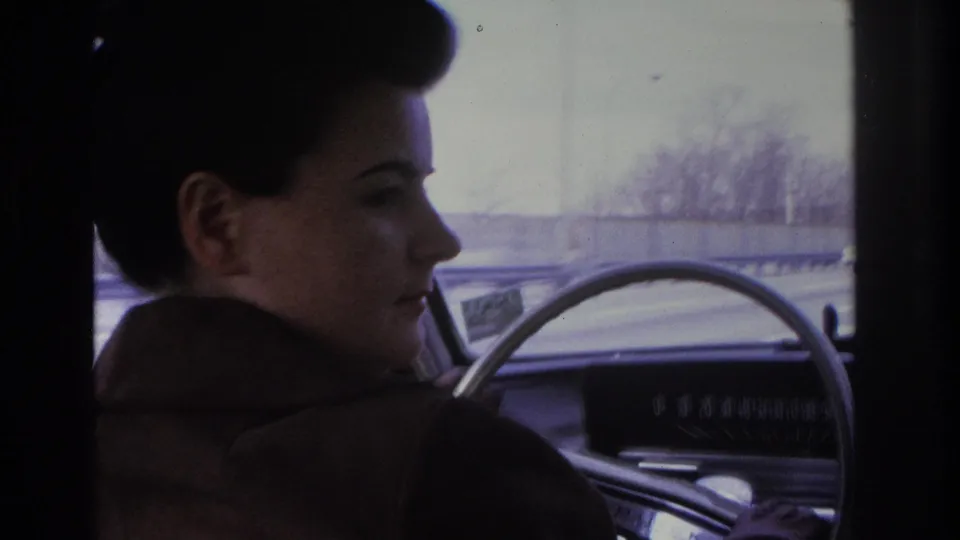 Three-quarter profile of a woman with dark styled hair and brown coat at the wheel; dashboard and overpass structure visible through windshield, mid-1960s.
