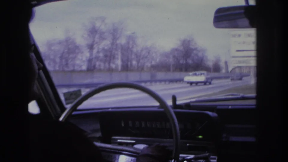 Interior shot from back seat: dashboard and steering wheel of a mid-1960s American car; multi-lane highway with passing trucks, bare trees, and partial highway sign through windshield.