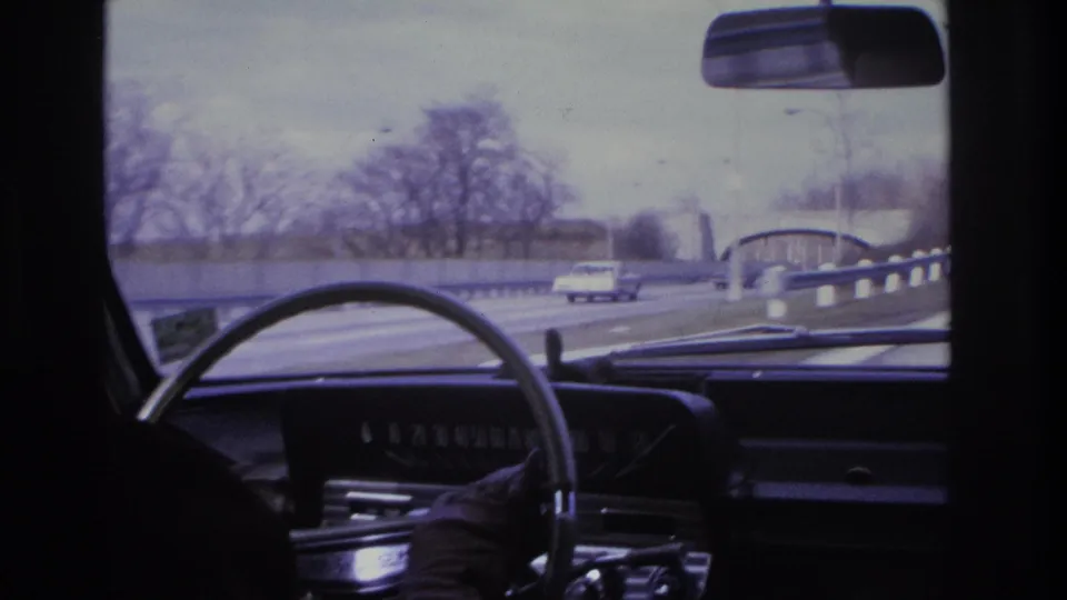 Interior dashboard shot of a mid-1960s American car; a highway bridge or overpass is visible in the middle distance through the windshield, with passing vehicles and bare trees.