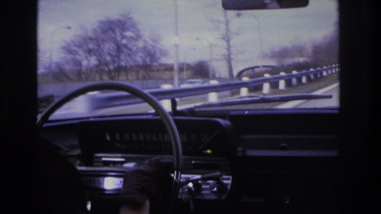 Driver's-eye view inside what appears to be a mid-1960s American car on a highway, possibly in New Jersey circa 1966, with dark instrument panel, large steering wheel, and bare winter trees.