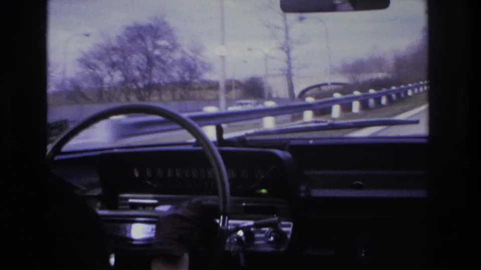 Driver's-eye view inside what appears to be a mid-1960s American car on a highway, possibly in New Jersey circa 1966, with dark instrument panel, large steering wheel, and bare winter trees.