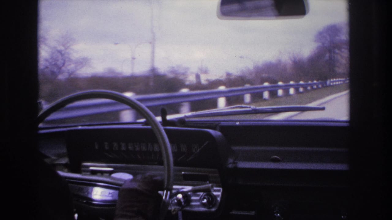 Driver's perspective in what appears to be a 1960s American car, showing the dark instrument panel and steering wheel with a guardrail-lined highway and utility poles visible ahead, circa 1966.